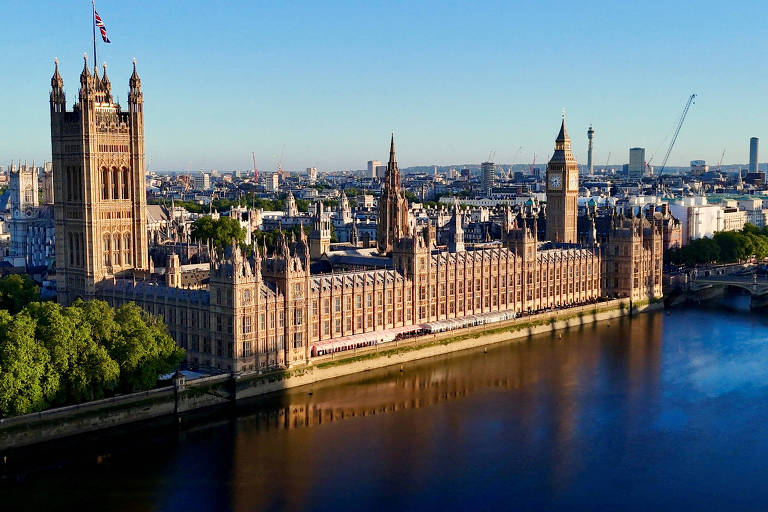 A imagem captura uma vista aérea do icônico Palácio de Westminster, sede do Parlamento do Reino Unido, banhado pela luz dourada do sol. O Rio Tâmisa serpenteia ao lado, refletindo a luz do dia, enquanto a cidade de Londres se estende ao fundo sob um céu claro e azul.