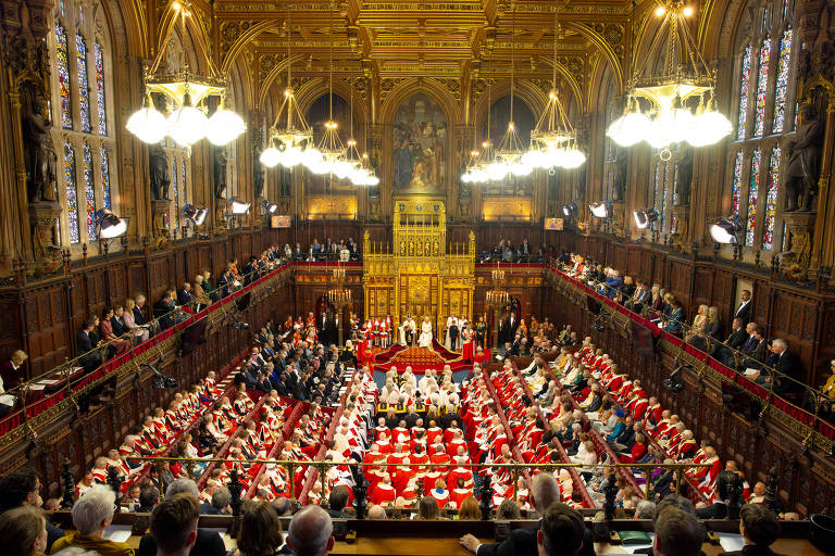 A imagem captura uma cerimônia formal na Câmara dos Lordes, com membros vestidos em trajes cerimoniais. É possível ver a arquitetura gótica ornamentada do interior e o uso de vermelho intenso nos assentos e trajes.