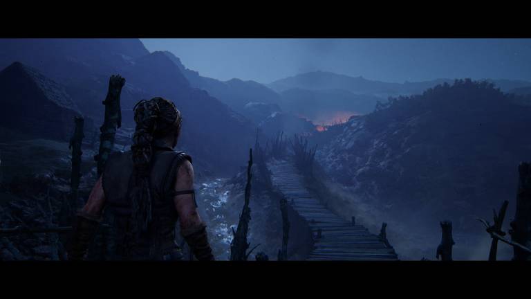 Um personagem de videogame, armado e com uma mochila, observa um vale montanhoso ao anoitecer. A luz suave do crepúsculo banha a paisagem, sugerindo um momento de calma