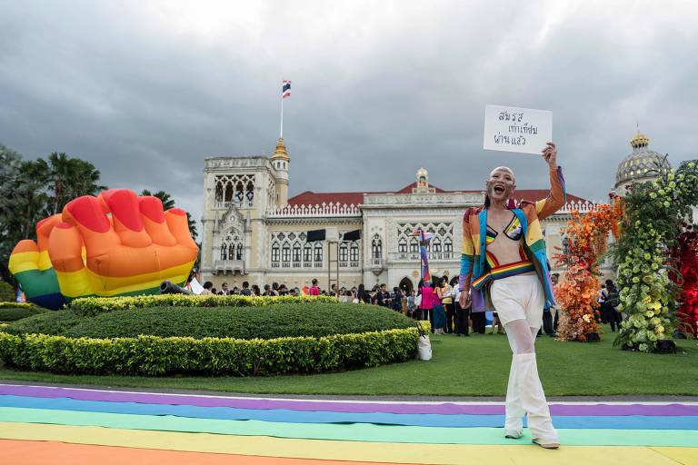 Uma pessoa exibe um cartaz e sorri amplamente enquanto participa de um evento colorido, com um caminho adornado com as cores do arco-íris e um inflável de mão em gesto de paz ao fundo, diante de um edifício histórico ornamentado.