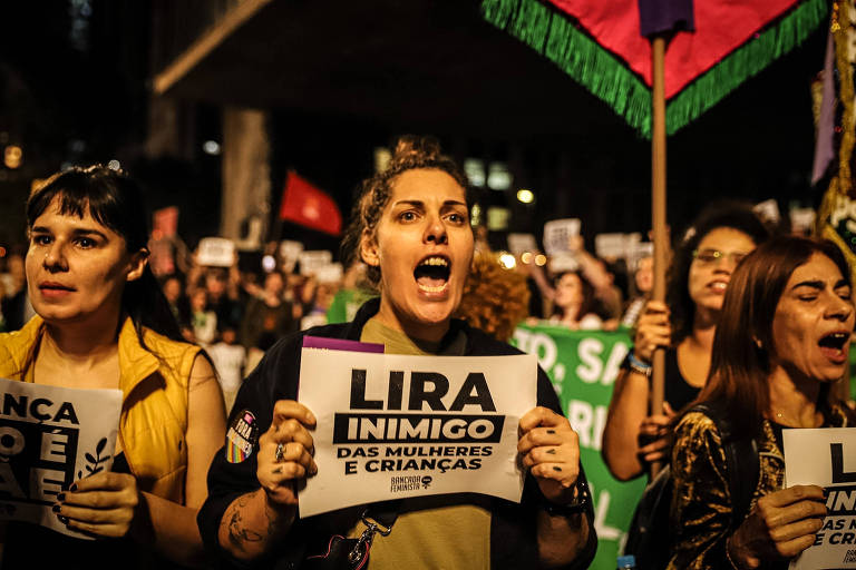 Imagem de um protesto noturno com várias mulheres segurando cartazes. No centro, uma mulher segura um cartaz que diz "Lira inimigo das mulheres e crianças". Outras mulheres ao redor também seguram cartazes e bandeiras, e parecem estar gritando