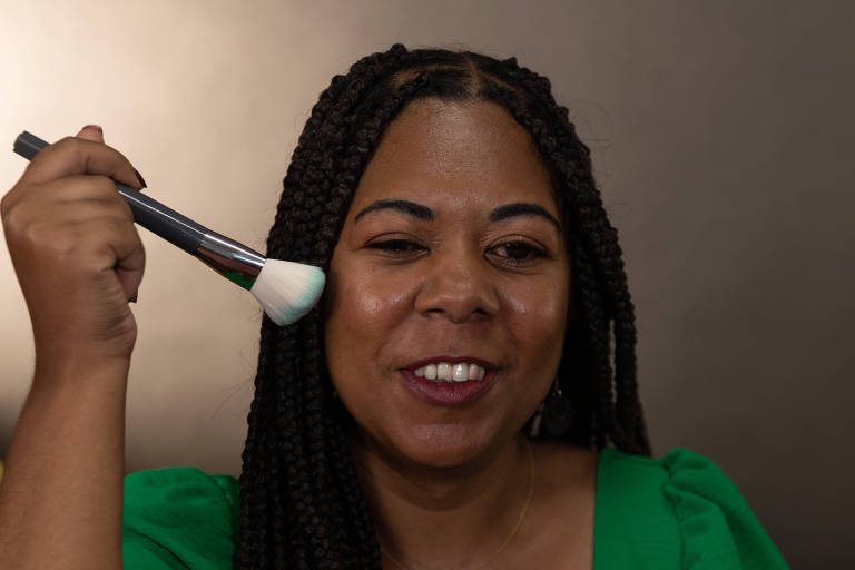 Uma mulher negra de tranças veste blusa verde e segura um pincel próximo ao rosto. 
