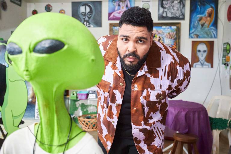 Em foto colorida,homem posa perto de uma figura de ET