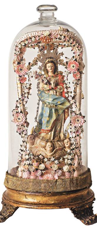 Uma delicada escultura religiosa, representando uma figura feminina possivelmente santa ou virgem, é protegida sob uma redoma de vidro. A figura central está adornada com uma vestimenta azul e dourada e é cercada por anjos e flores intricadamente detalhadas