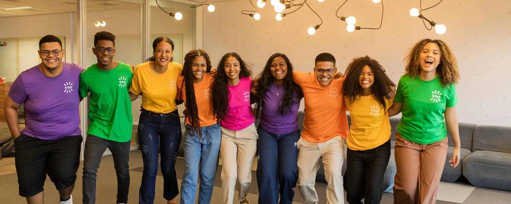 Um grupo diversificado de jovens adultos sorri e posa em um ambiente de escritório, vestindo camisetas coloridas. A iluminação e o design moderno do local complementam a atmosfera alegre e colaborativa que o grupo exibe.