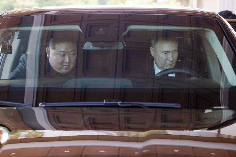 Dois homens são vistos através da janela de um veículo. O homem no banco do passageiro, vestindo um terno escuro, olha atentamente para o outro, que está ao volante e também vestido formalmente.