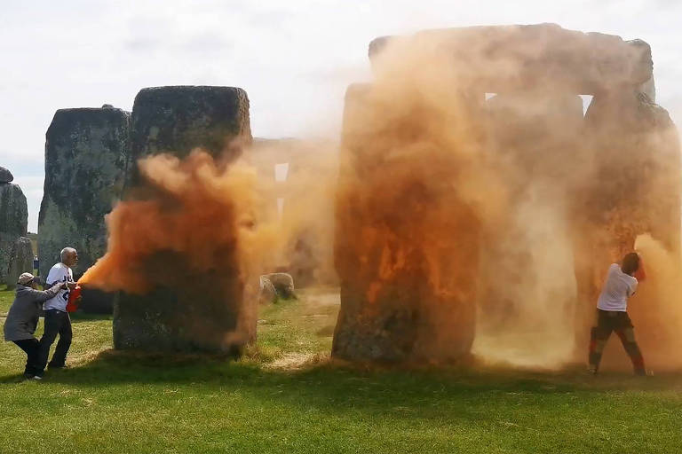 Três pessoas perto das pedras de Stonehenge; há tinta laranja voando no ar, perto das rochas