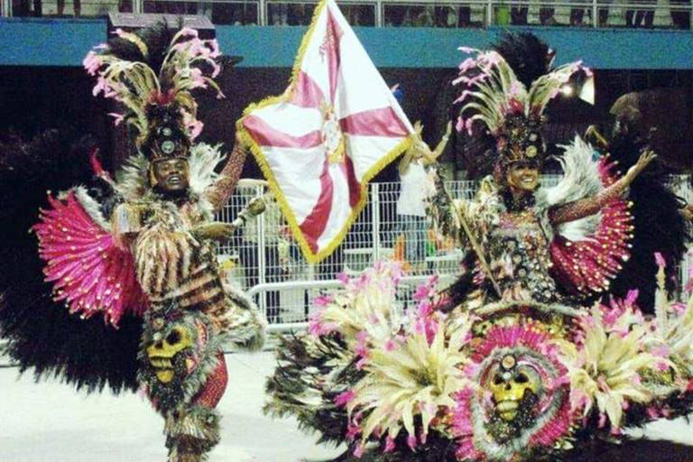 A imagem captura a essência vibrante do Carnaval, mostrando dois participantes em trajes extravagantes e coloridos, adornados com penas e brilhos. Eles parecem estar em um desfile, com um deles carregando uma bandeira, ambos exibindo sorrisos radiantes que refletem a alegria e o espírito festivo do evento.