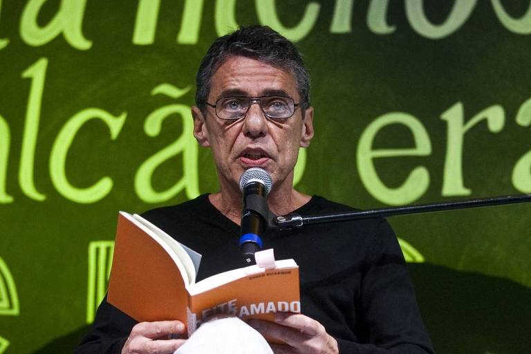 homem branco de óculos lê livro laranja em microfone contra fundo verde com letras