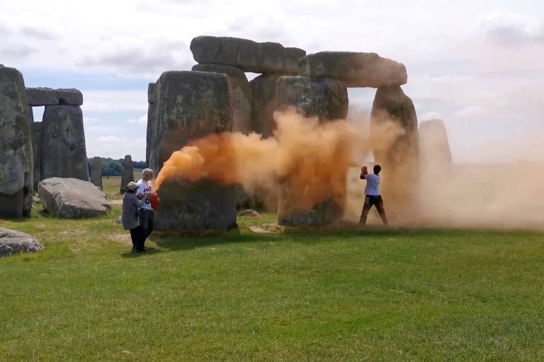 Duas pessoas jogam tinta laranja em pedras em uma planície; ao lado de uma das pessoas, está uma outra que parece estar puxando um dos manifestantes para longe da pedra, tentando impedir a ação