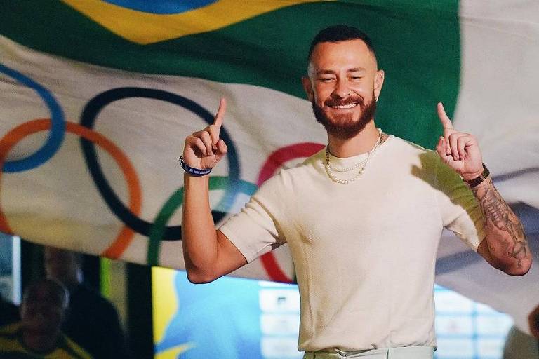 Um homem sorridente posa diante de uma bandeira do Brasil, que ocupa o fundo da imagem. Ele está fazendo um gesto com as mãos, apontando para cima com os dedos indicadores, e exibe tatuagens nos braços. Atrás dele, a bandeira é complementada por um símbolo olímpico.