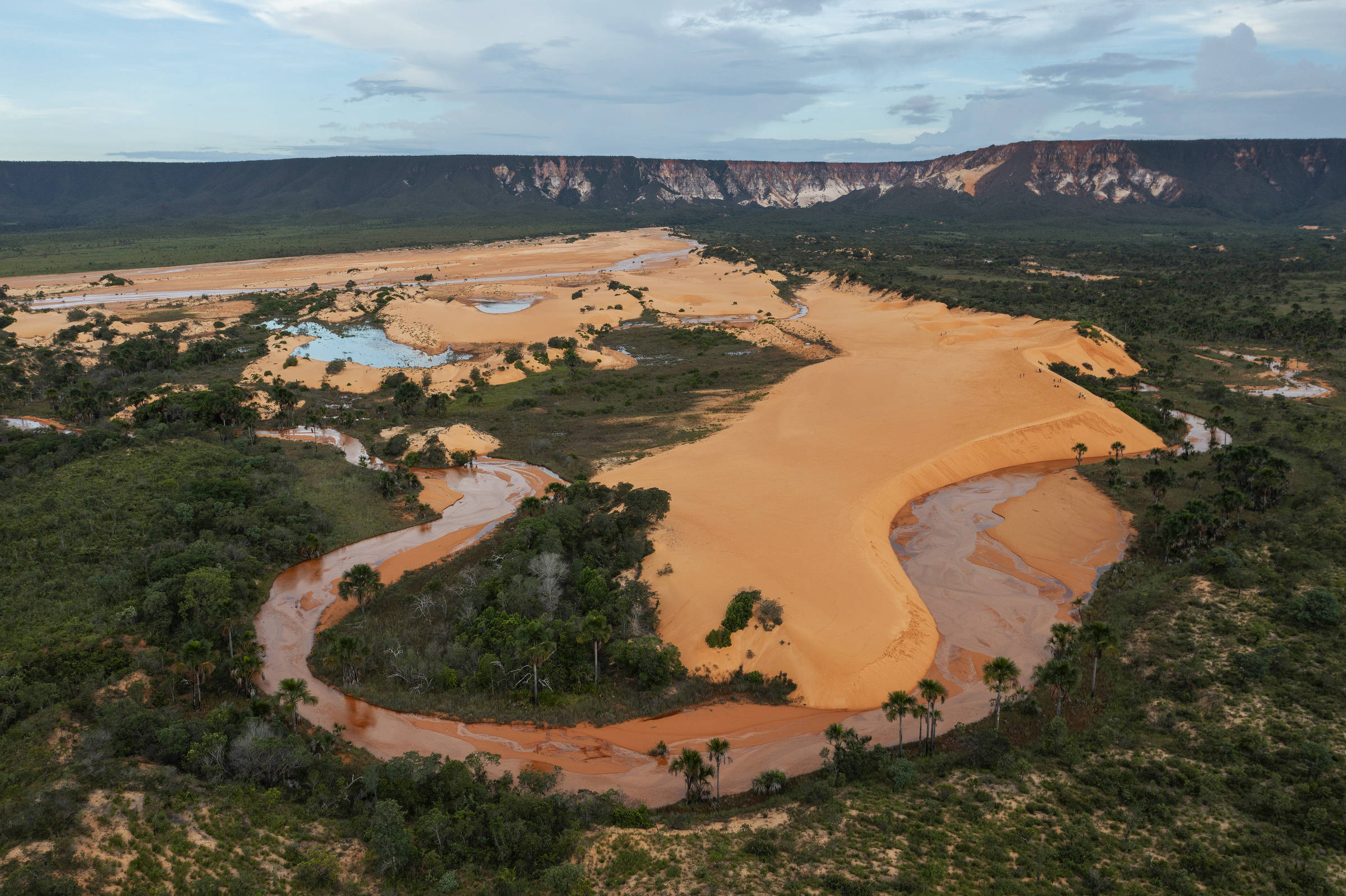 Um rio sinuoso se entrelaça com bancos de areia dourada, cercado por uma vegetação densa e falésias ao fundo