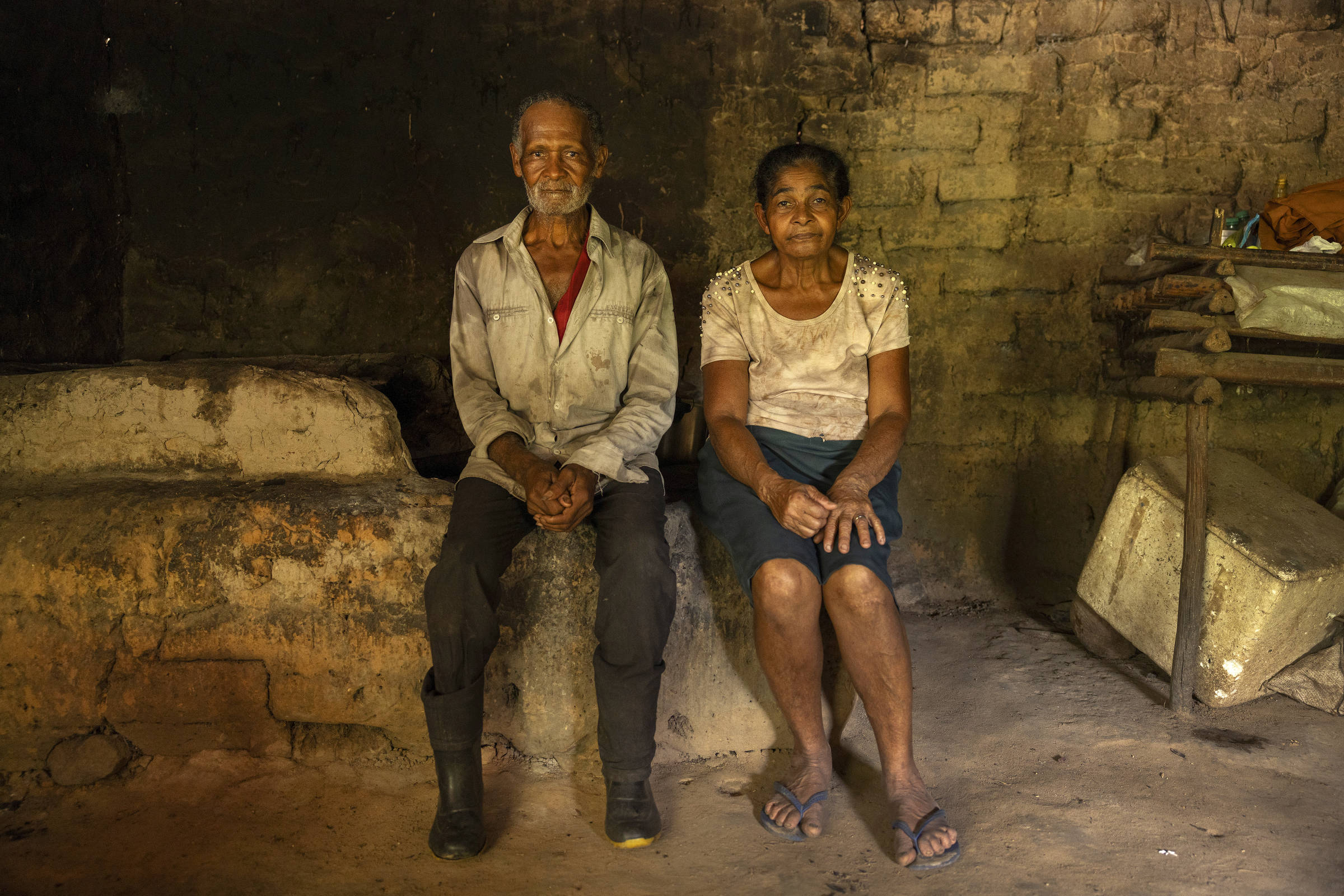 Um homem e uma mulher idosos, sentados lado a lado em um banco rústico dentro de uma casa simples com paredes de barro