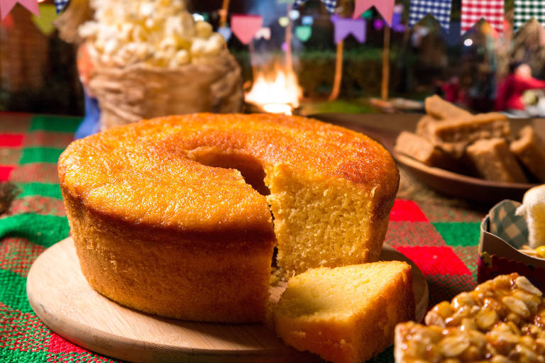 Em primeiro plano, um bolo de milho repousa sobre uma tábua de madeira, com uma fatia cortada. Ao fundo, uma mesa de festa junina é adornada com comidas típicas e decoração colorida, incluindo bandeirinhas e uma fogueira ao longe