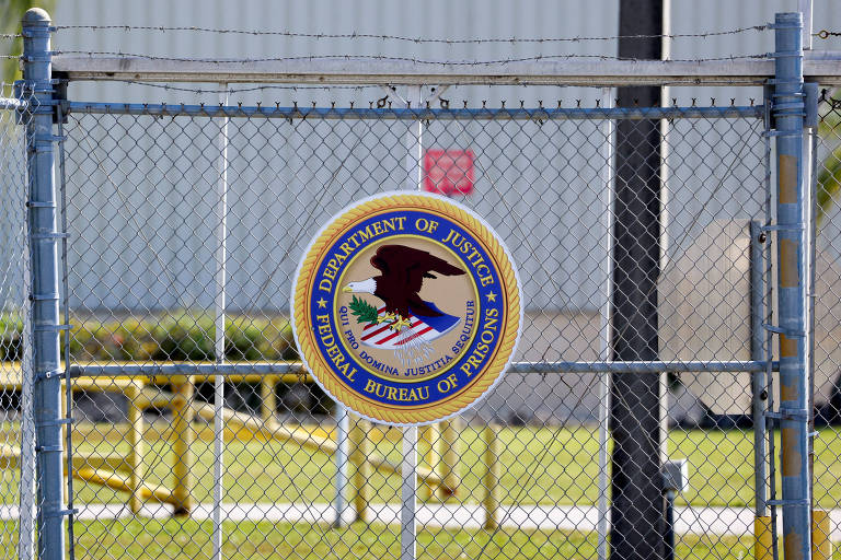 O selo da agência federal de prisões é visto aqui fixado em uma cerca de arame. O selo é claramente visível, com a águia americana no centro, enquanto o fundo desfocado sugere um local protegido ou restrito.