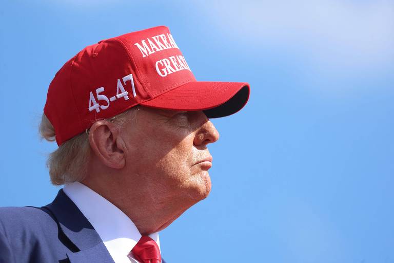 Donald Trump, com expressão séria, olha para o horizonte, vestindo um boné vermelho contra um céu azul claro