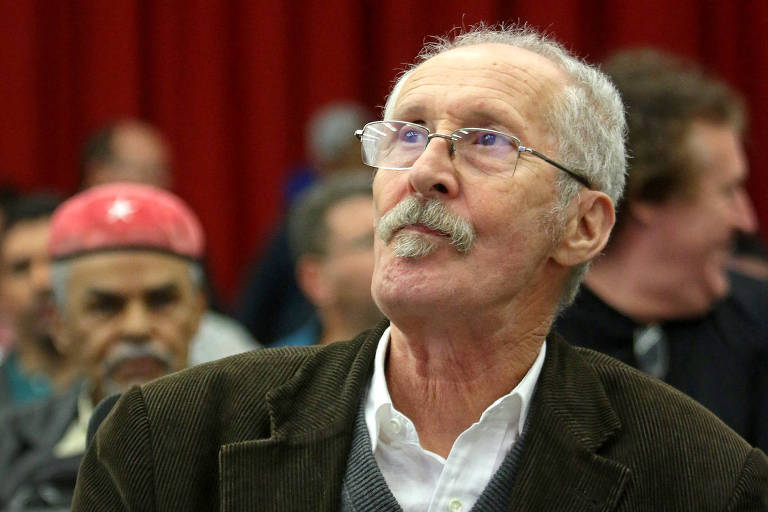 Um homem mais velho com bigode e óculos, vestindo um blazer marrom e uma camisa branca, olha para cima com uma expressão pensativa, em frente a uma cortina vermelha, enquanto outras pessoas parecem desfocadas ao fundo.