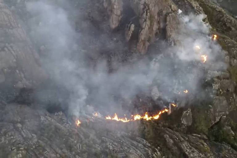 A imagem mostra uma encosta rochosa com sinais de um incêndio ativo, onde linhas de fogo podem ser vistas serpenteando entre as pedras. Uma densa fumaça cinza sobe da área queimada, indicando a presença de um incêndio florestal ou de vegetação.