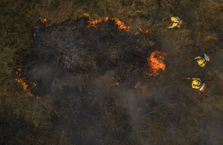 foto aérea na vertical mostra agentes no solo usando equipamentos para conter fogo. há uma área queimada e o fogo se espalha para fora dela, em direção à vegetação 