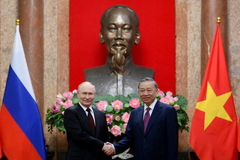 Dois homens de terno apertam as mãos em frente a um busto de bronze e uma parede com cortinas vermelhas, simbolizando um momento de diplomacia e cooperação internacional. As bandeiras da Rússia e do Vietnã estão posicionadas ao fundo, indicando as nações representadas.