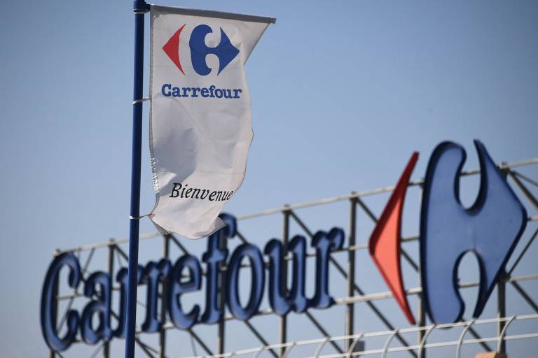 A imagem mostra duas bandeiras ao vento, uma com o logotipo do Carrefour e outra com o nome "Carrefour" escrito em letras grandes, ambas contra um céu azul claro. A bandeira com o logotipo apresenta o design com duas setas azul e vermelha apontando para um "C" central.