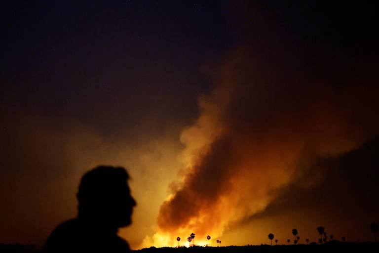 A imagem captura a silhueta de uma pessoa observando de longe um grande incêndio que ilumina o céu noturno. As chamas alaranjadas e a fumaça densa contrastam com o céu, enquanto algumas árvores se destacam contra o brilho do fogo.