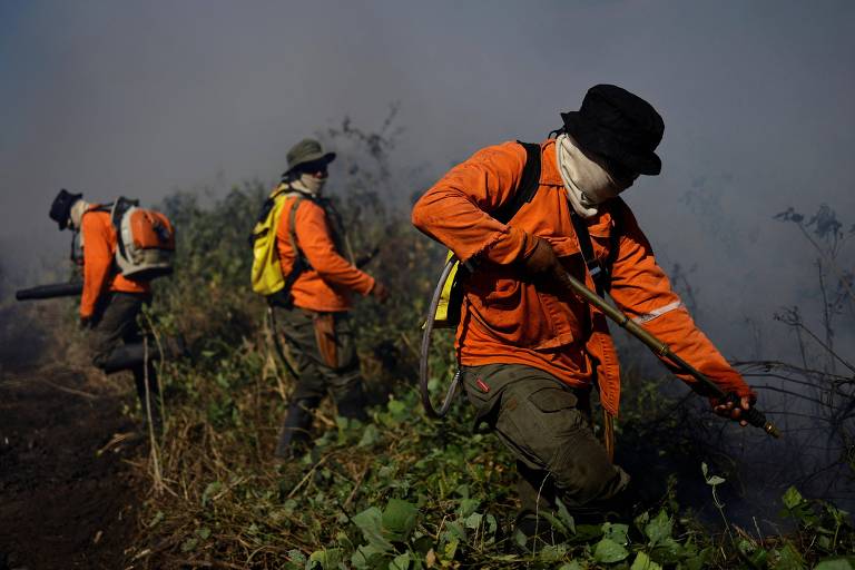 Vestidos com uniformes laranja e equipamentos de proteção, brigadistas combatem incêndio enquanto uma densa fumaça se espalha ao fundo