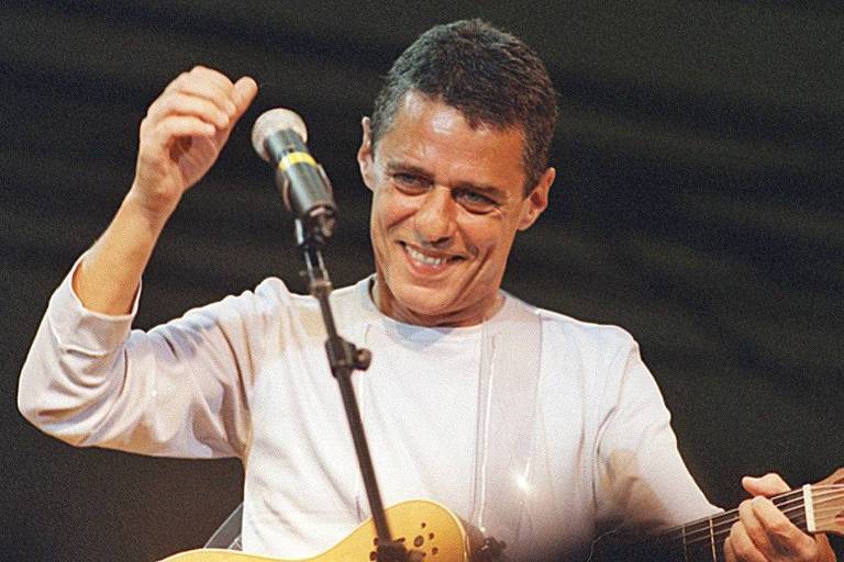 Homem branco de camisa branca sorri diante de microfone enquanto segura violão e ergue o braço em saudação