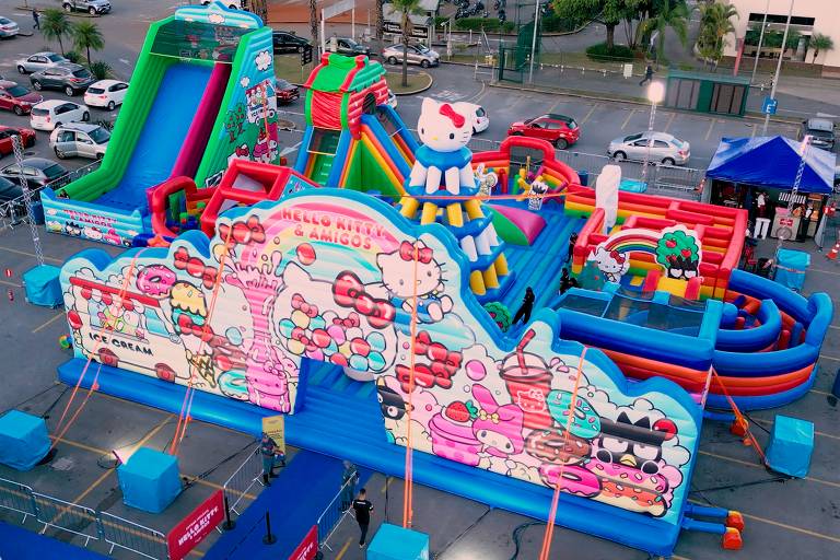 A imagem mostra um grande parque inflável temático da Hello Kitty, localizado em um estacionamento. O parque é colorido e possui várias áreas de brincadeiras, incluindo escorregadores, obstáculos e uma grande estrutura central com a figura da Hello Kitty no topo. A entrada do parque tem a inscrição 'HELLO KITTY & AMIGOS' e é decorada com desenhos de sorvetes, doces e personagens da Hello Kitty. Há várias pessoas ao redor do parque, e carros estacionados ao fundo.