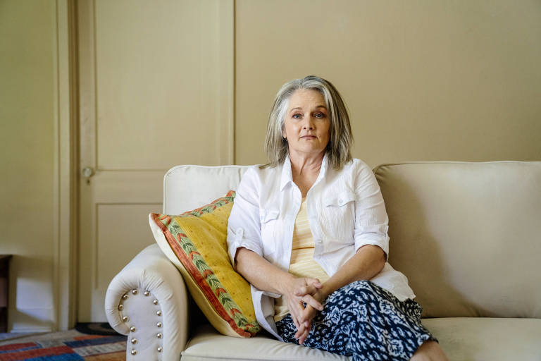 Uma mulher de cabelos grisalhos está sentada em um sofá bege em uma sala de estar. Ela veste uma camisa branca e uma saia com padrão azul e branco. Há uma almofada amarela com detalhes coloridos ao seu lado