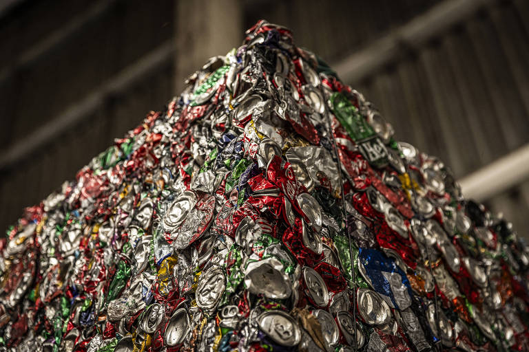 A imagem mostra uma pilha de latas de alumínio comprimidas. As latas estão amassadas e misturadas, formando um bloco compacto. Há uma variedade de cores visíveis nas latas, incluindo vermelho, verde, azul e prata
