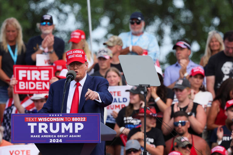 Imagem de um comício político onde Trump, de terno azul e gravata vermelha, usando um boné vermelho, está falando em um púlpito. Atrás dele, há uma multidão de pessoas, algumas segurando cartazes com mensagens como 'FIRE BIDEN' e 'VOTE'.