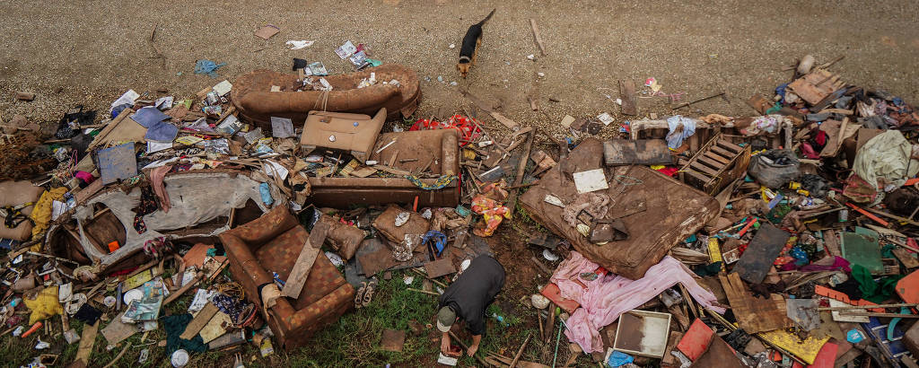 Foto aérea mostra homem em meio a entulho de lixo, móveis quebrados e objetos descartados; um cachorro se aproxima