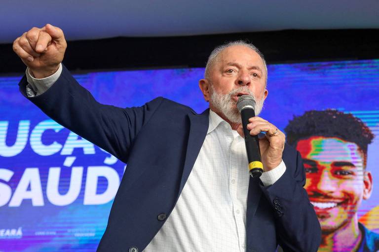 Lula, homem de barba grisalha e cabelo curto, está discursando com um microfone em uma mão e gesticulando com a outra. Ele veste um terno azul escuro e uma camisa branca. Ao fundo, há um painel com as palavras 'EDUCAÇÃO' e 'SAÚDE' parcialmente visíveis, além da imagem de um jovem sorridente.
