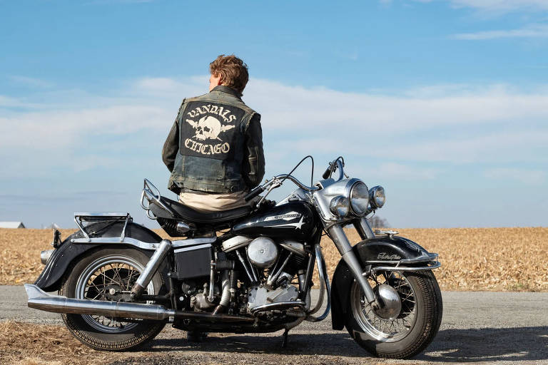 Uma pessoa sentada em uma motocicleta preta,está de costas, vestindo uma jaqueta de couro com as palavras 'Vandals Chicago'