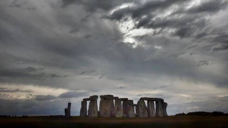 A imagem mostra o monumento de Stonehenge, composto por grandes pedras dispostas em círculo, sob um céu nublado e escuro. O horizonte é baixo, destacando as pedras contra o céu dramático.