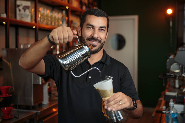 Homem pardo de camisa preta despeja água quente sobre um filtro de café. Ele está sorrindo