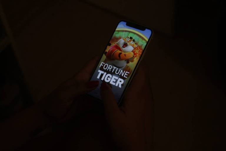 A imagem mostra uma pessoa segurando um celular com a tela exibindo o jogo 'Fortune Tiger'. A tela do jogo mostra um tigre animado segurando um objeto vermelho com caracteres chineses. A mão da pessoa tem unhas pintadas de vermelho.