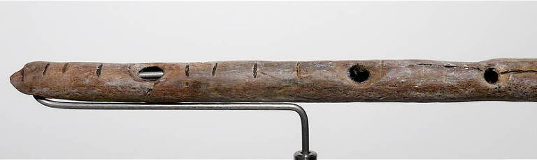 A imagem mostra uma flauta de madeira antiga, com uma aparência rústica e desgastada. A flauta possui três buracos visíveis. A madeira parece envelhecida e apresenta algumas rachaduras e marcas de uso.