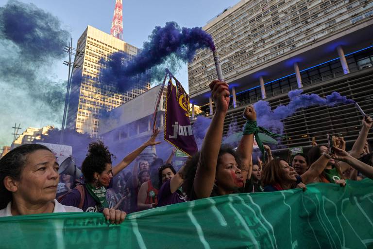 Mulheres protestam segurando faixa verde e sinalizadores que soltam fumaça azul e roxa; atrás delas estão prédios altos