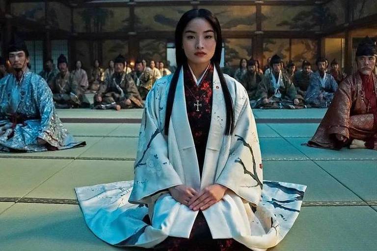 Uma das cenas de "Xógum" em que Mariko, uma mulher jovem, oriental, de longos cabelos escuros, está vestida de branco, sentada em um tatame. No segundo plano, dois homens, um de cada lado e, mais ao fundo, outras fileiras de pessoas sentadas.