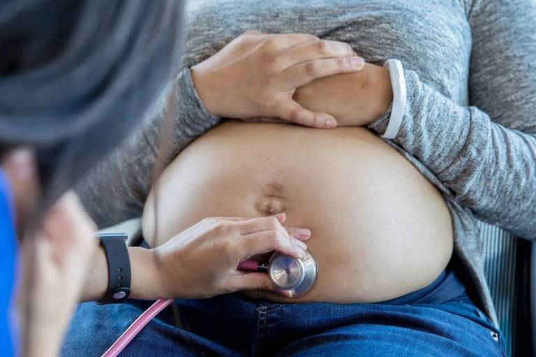 Foto mostra uma médica, de pele clara, colocando estetoscópio em barriga de mulher grávida também de pele clara.