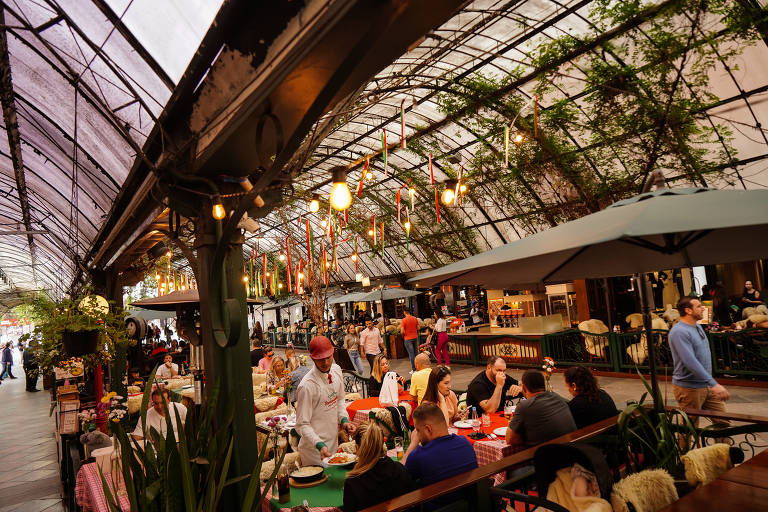 A imagem mostra um mercado interno com um teto de vidro, onde várias pessoas estão sentadas em mesas. O ambiente é decorado com plantas e há iluminação pendente. Algumas pessoas estão interagindo, enquanto outras estão concentradas em suas refeições. O espaço parece ser animado e acolhedor.