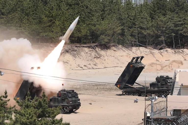Numa praia, um míssil branco é lançado de um veículo militar de cor verde, deixando rastro de fumaça
