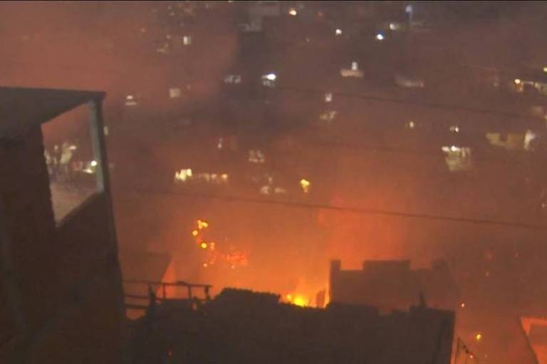 A imagem mostra um incêndio em uma favela em São Paulo durante a noite. O fogo é visível em várias áreas, com chamas intensas e muita fumaça.