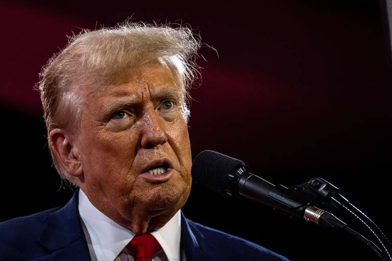 A imagem mostra um homem de cabelos loiros e pele clara falando ao microfone. Ele está vestindo um terno escuro, camisa branca e gravata vermelha. O fundo é escuro com tons de vermelho.