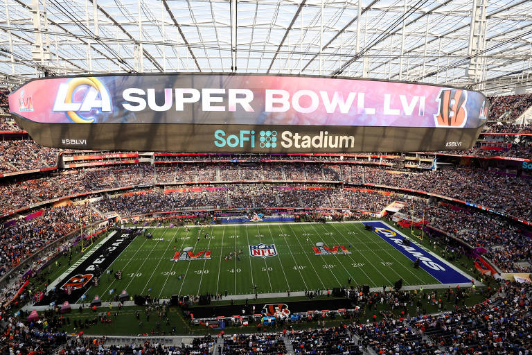 Imagem de um estádio de futebol americano lotado durante o Super Bowl LVI. O telão exibe 'SUPER BOWL LVI' com os logotipos dos Los Angeles Rams e dos Cincinnati Bengals. Abaixo do telão, está escrito 'SoFi Stadium'. O campo está preparado para o jogo, com as marcações e logotipos dos times visíveis.