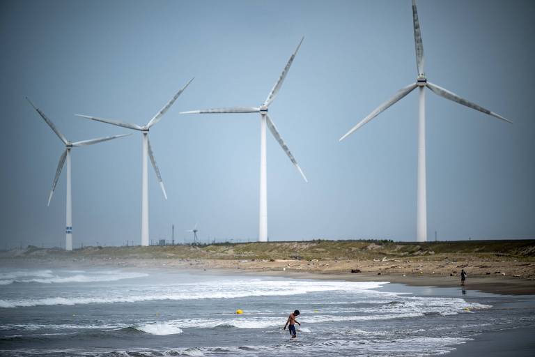 A imagem mostra quatro grandes turbinas eólicas instaladas em uma área costeira, próximas ao mar. No primeiro plano, duas pessoas estão na água, uma mais próxima da câmera e outra mais distante. O céu está claro e a água do mar está agitada com ondas.