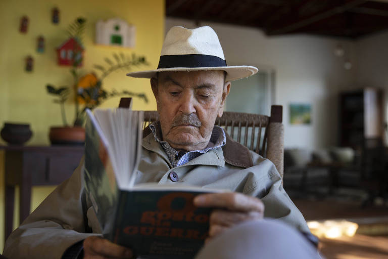 Homem branco de barba grisalha e chapéu lê livro sentado