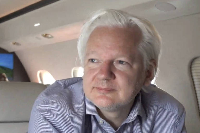 A imagem mostra um homem de cabelos brancos sentado em um avião. Ele está vestindo uma camisa azul clara e olhando para o lado. O interior do avião é visível ao fundo, com janelas e assentos.